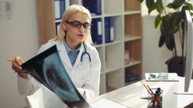 Gözlüklü çekici doktor hasta hastaya röntgendeki görüntüyü açıklıyor. Sarışın Ukraynalı dost canlısı kadın doktorun ofisinde oturmuş konuşuyor..