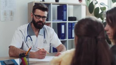 Gözlüklü ve beyaz önlüklü kendinden emin bir çocuk doktoru fiziksel muayeneden önce kızla konuşuyor ve muayenehanedeki belirtileri soruyor..