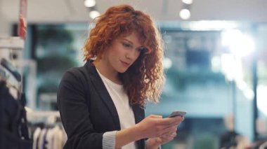 Kırmızı kıvırcık saçlı, gülümseyen genç bir kadın telefon tasarımcısı kızı kullanarak dükkânda dikiliyor. Güzel, çekici, hoş, kıvırcık saçlı, genç, yavaş çekimde çekilmiş bir portre.