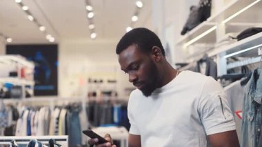 Yakışıklı Afro-Amerikan genç adam dükkandaki telefon mesajı standını kullanıyor. Giysileri seçiyor. Mutlu siyah telefon portresi.
