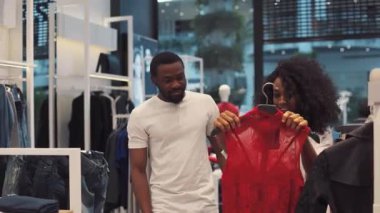 Şirin Afro-Amerikan çifti kıyafet mağazasından kıyafet seçiyor gülümseyen aile kadını moda alışverişi yapan adam dükkan sahibi, kamera erkek arkadaşına bakıyor genç kız arkadaş.