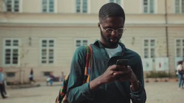 Genç gülümseyen Afrikalı Amerikalı gözlüklü adam şehirde telefon kullanıyor iş adamları meşgul insanlar modern siyah kararlı yakışıklı yaşam tarzı hırslı cep telefonları