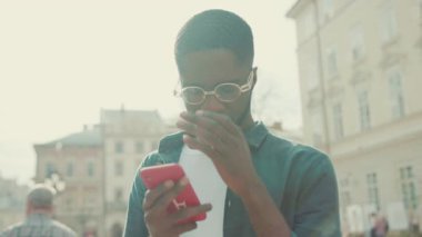 Gözlüklü üzgün Afrikalı Amerikalı genç adam şehir portresindeki telefon kulübesini kullanıyor. Günbatımı teknolojisi. İş adamları meşgul.