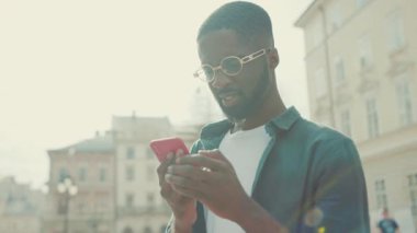 Gözlüklü Afro-Amerikalı genç adam şehirde telefon kulübesi kullanıyor mutlu gülüş teknolojisi işadamları meşgul modern siyah portre yakışıklı yaşam tarzı akıllı telefon hırslı mobil gün batımı