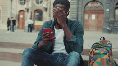 Gözlüklü üzgün Afrikalı Amerikalı genç adam şehirde oturmuş ciddi sinirli teknoloji insanları cezbediyor modern siyah yakışıklı akıllı telefon hırslı mobil gün batımı