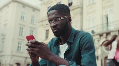 Kapalı kapılar ardında gözlüklü Afro-Amerikan genç adam şehirde oturmuş gülümse teknolojisi meşgul insanlar modern siyah yakışıklı akıllı telefon hırslı mobil günbatımı ağır çekim