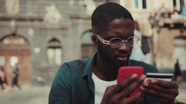 Gözlüklü Afrikalı Amerikalı genç adam telefon kartı kullanıyor. İnternetten alışveriş teknolojisi alıyor. Meşgul portresi.