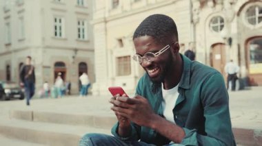 Çekici, mutlu yüzlü, gözlüklü, gözlüklü, şehirde oturan, gülümseyen, teknolojili, modern, yakışıklı, akıllı telefonlu, hırslı, mobil günbatımı ağır çekimde portresi olan Afro-Amerikan genç adam.