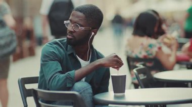 Ciddi Afro-Amerikalı gözlüklü, müzik dinleyen, kulaklık takan, kahve içen, moda yüzlü, siyah kulaklıklı, mutlu, ağır çekimde kulaklık takan bir adam.