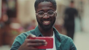 Gözlüklü genç Afrikalı Amerikalı adam akıllı telefon kullan. Bir kafede oturup gülümse. Kahve iç, moda teknolojisi iç.