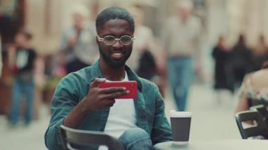 Kapatın gözlüklü genç Afrikalı Amerikalı adam. Akıllı telefon kullanın. Bir kafede oturup gülümseyin.