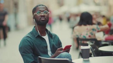 Gözlüklü Afro-Amerikalı bir adam kulaklık takıyor. Akıllı telefon kullanıyor. Bir kafede oturuyor, kahve içiyor.