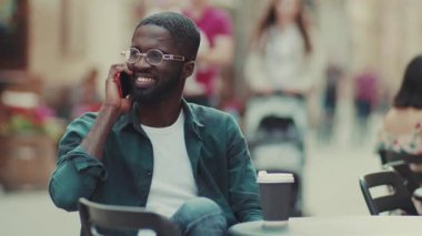 Gözlüklü genç Afrikalı Amerikalı adam akıllı telefonuyla konuşuyor. Caddedeki bir kafede oturuyor. Kahve içiyor, moda, siyah kulaklık takıyor.