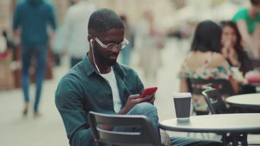 Gülümseyen, gözlüklü, kulaklık takan, akıllı telefon kullanan, kahve içen, kahve içen, siyah kulaklıklı, siyah kulaklıklı, mutlu genç bir adam.