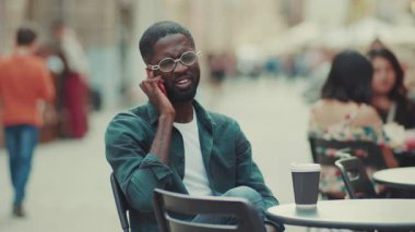 Gözlüklü mutlu Afrikalı genç adam akıllı telefonuyla konuşuyor. Bir kafede oturup kahve içiyor. Moda, siyah kulaklıklar, çekici, yakışıklı, mutlu, yavaş çekim.
