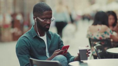 Gözlüklü genç Afrikalı Amerikalı adam kulaklık takıyor. Akıllı telefon kullanıyor. Bir kafede oturuyor, kahve içiyor.