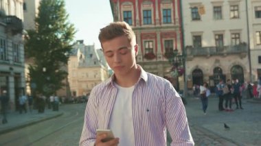 Portre genç mutlu adam sokakta yürür etrafına bak gülümse güneş ışığı sevimli iş mobil aygıtı şehir dışında akıllı telefon teknolojisi yakışıklı dokunmatik ekranı yavaş çekim kapat