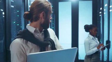 Adanmış ve odaklanmış bir takım üyesi sunucularla dolu modern bir veri merkezinde özenle bir dizüstü bilgisayar kullanıyor.