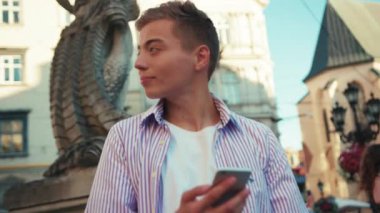 Elinde cep telefonu olan gülümseyen hippi çocuk şehir merkezinde mutlu bir şekilde dikil. İnternetin dışında genç ve yakışıklı bir öğrenci caddesi tut. Akıllı telefon teknolojisi cep telefonu ağı.