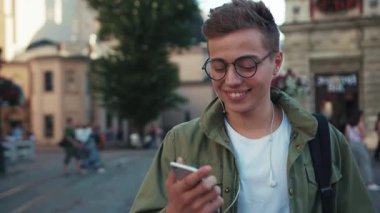 Gözlüklü bir portre genç adam telefon kullanıyor kulaklıkla müzik dinliyor mutlu bir cep telefonu ışığı yürüyüşü aleti açık havada akıllı telefon teknolojisi yakışıklı hippi yavaş çekim kapat