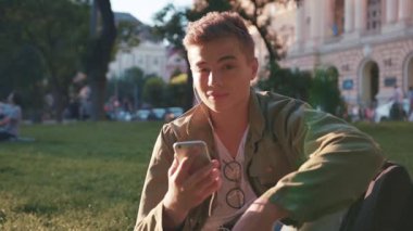 Parkta oturmuş akıllı telefon kullanan gülümseyen genç adam kameraya bak. Gün batımı öğrencisi mutlu ol. Elinde yakışıklı mesajcı çocuk eli, ağır çekimde beyaz portre.