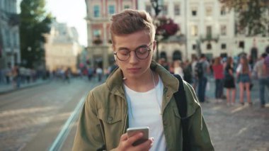 Kapalı yüzlü mutlu genç adam telefon kullan kulaklıkla müzik dinle cep telefonu gülümse cep telefonu yürü mobil cihaz açık havada akıllı telefon teknolojisi yakışıklı hippi yavaş çekim portresi