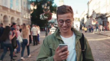 Gözlüklü hoş bir genç adam telefon kullanıyor kulaklıkla müzik dinliyor mutlu bir cep telefonu ışığı gezici cihaz dışarıda akıllı telefon teknolojisi yakışıklı hippi yavaş çekim portresini kapat