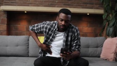 Genç ve çekici Afrikalı adam gri koltukta oturuyor, oturma odasında cep telefonu kullanıyor. Ekose gömlekli hoş bir adamın mideye dokunuşunun portresi. Keskin bir acı. Kapalı.