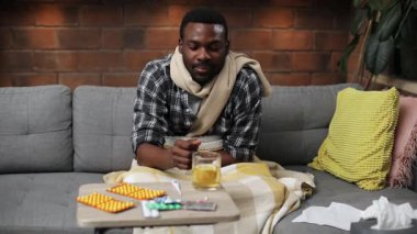 Bej eşarp takan, gri koltukta oturan, renkli yastıklı, öksüren Afrikalı genç bir adam. Çekici bir adam donuyor, çay içiyor, başka tarafa bakıyor. Oturma odası.