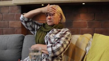 Kanepede, renkli yastıkları ve vücut ısısını ölçen yaşlı, beyaz bir kadın oturuyor. Büyükannem kameraya dikkatle bakıyor. - Eve. Oda.