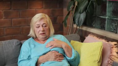 Slav, mavi kazaklı yaşlı kadın, yüreğine dokunuyor. Acı çeken büyükannenin kalp ağrısı portresi. İçeride. Yakın plan. Derin nefes al..