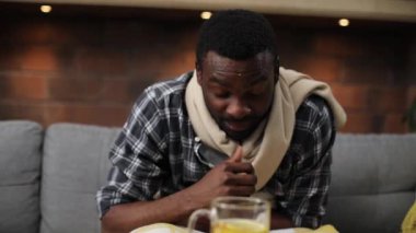 Kanepede oturan, öksüren, dondurucu soğuk içinde hasta Afrikalı bir adam. Sıcak çay içen genç bir adamın portresi. Yüksek ateş. Sıcaklık. Kapalı.