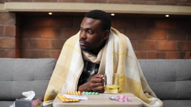 Ekose battaniyeli yakışıklı hasta genç adam renkli uyuşturucularla birlikte masanın yanındaki koltukta oturuyor. Çekici Afrikalı adamın portresi. Yakın plan. İçeride. Hastalık.