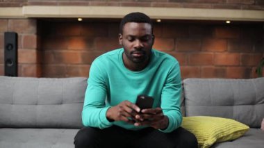 Yüzüne dokunan sakallı hoş bir Afrikalı. Elinde siyah akıllı telefon tutan naneli süveter giymiş hoş bir gencin portresi. İçeride. Oturma odası. Diş ağrısı..