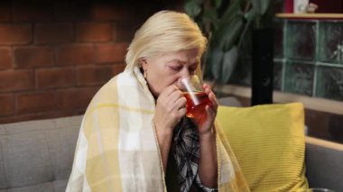 Kanepede oturan yaşlı kadın, elinde bir fincan çay tutuyordu. Kameraya bakan üzgün, beyaz kadın portresi. İçeride. Sıcaklık. Hastalık..