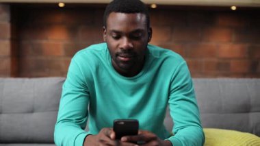 Nane kazaklı, odaklanmış Afrikalı bir adamın portresi elinde akıllı telefonu tutarak ekrana dikkatle bakıyor. Baş ağrısı. Büyük ev odası. Sıcaklık.