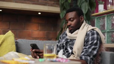 Kanepede battaniyenin altında yatan yakışıklı Afrikalı adam. Tatlı bir genç adamın portresi, dondurucu soğuk, sıcak çay içen, akıllı telefon kullanan. İçeride. Oturma odası.