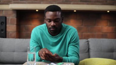 Elinde ilaçlarla kanepede oturan, haplardan konuşan, kameraya dikkatle bakan yakışıklı bir Afrikalı. Yakın plan. Oturma odası. Kapalı.