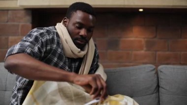 Rahat kıyafetler giyen yakışıklı genç adam kanepede oturuyor ve vücut ısısını ölçüyordu. Yakışıklı, hasta, Afrikalı bir adamın portresi. Kapalı.