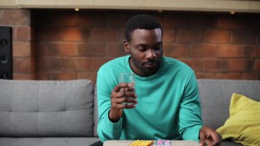 Kanepede oturmuş elinde haplarla naneli süveter giyen yakışıklı bir Afrikalı. İyi bir genç adam doğrudan kameraya bakıyor, gülümsüyor. - Eve. Kapalı.