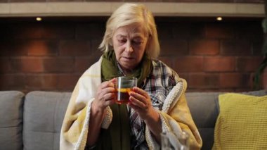 Kafkasyalı hasta kadın kanepede oturup limonlu sıcak çay içiyor. Düşünceli mutsuz büyükanne başka tarafa bakıyor. İçeride. Sıcaklık. Ev.