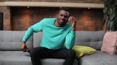 Kanepede oturan yakışıklı genç adam renkli yastıklarla, elinde akıllı telefonu tutarak ekrana bakıyor. Kafaya dokunan Afrikalı adamın portresi. Migren..