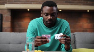 Çekici Afrikalı odaklı genç adam haplarla dolu masanın yanında oturuyor. İlaçlara bakıp seçen mutsuz bir adamın portresi. İçeride. Yakın plan..