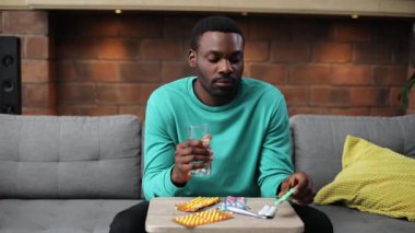 Sakallı hasta Afrikalı adamın portresi. Spor elbiseli çekici bir genç adam kanepeye oturmuş, ilaçlara bakıyor, elinde bir bardak su tutuyor. Tedavi.
