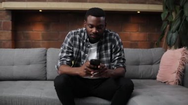 Kanepede oturan, akıllı telefon kullanan, gündelik kıyafetler giyen hoş bir adamın portresi. Genç, hoş, Afrikalı bir adam arkadaşlarıyla sohbet ediyor, kafaya dokunuyor. Korkunç migren..