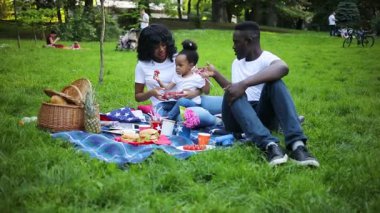 Yeşil parkta piknik battaniyesinde oturan hoş bir Afrikalı aile. Anne ve babası çok tatlı bir çocuk taşıyor, anne kızına çilek veriyor. Yaz zamanı. Açık havada. Yaşam biçimi.