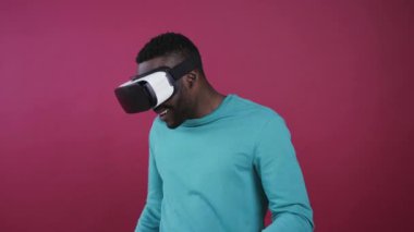 Parlak mavi gömlekli yakışıklı Afro-Amerikalı adam sanal gerçeklik kulaklığını takıyor, etrafa neşeyle bakıyor, kaçıyor, kalkıyor ve renkli arka planda gülümsüyor. Modern teknolojiler. Erkek portresi