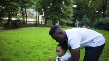 Kızlarının elini tutan beyaz tişörtlü çekici Afrikalı genç bir adam. Baba ve kızı yeşil çimlerde yürüyor. Açık havada. Yaz zamanı. Gündüz.