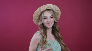 Çiçekli elbiseli ve şirin şapkalı güzel bir kadın kameraya döner, mutlu bir şekilde gülümser, saçlarıyla baştan çıkarıcı bir şekilde oynar, pembe bir arka planda döner. Eğlenmek, iyi hissetmek, olumlu duygular. Kadın portresi