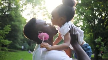 Afrikalı genç bir baba küçük tatlı bir kızı elinde tutuyor. Baba sevimli kızını öpüyor. Güneş ışığı. Yaz zamanı. Hafta sonu. Açık havada. Gündüz.